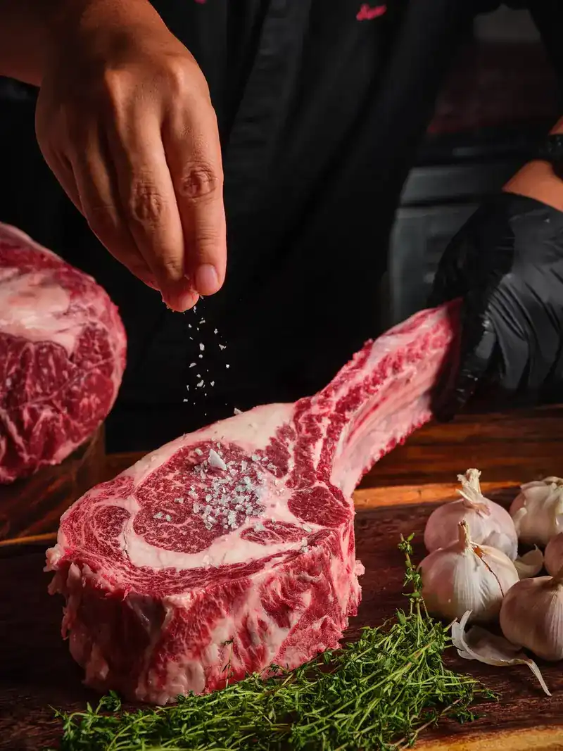A hand scattering salt flakes over a raw long-bone rib steak on a wooden block
