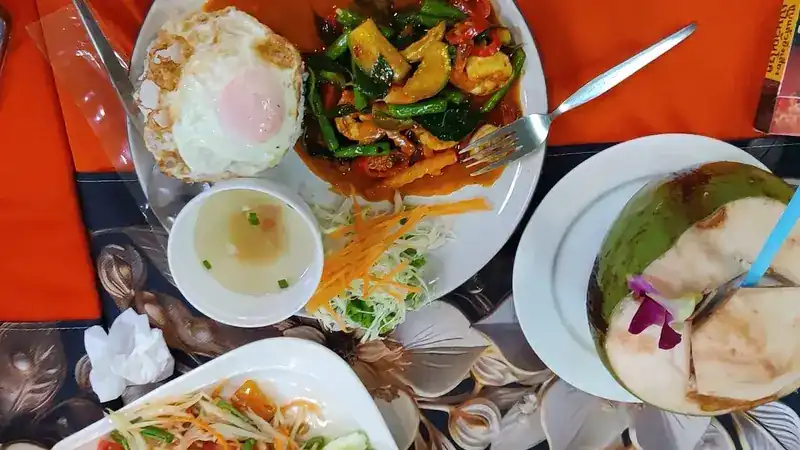 Seen from above: a stir-fry with a fried egg, a bowl of soup, salad and a fresh coconut on an orange tablecloth