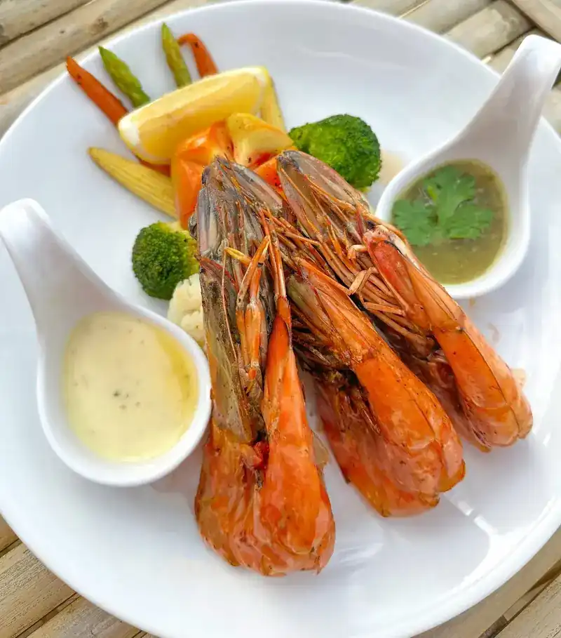 Large prawns on a white plate with vegetables and two small spoons of dipping sauce.