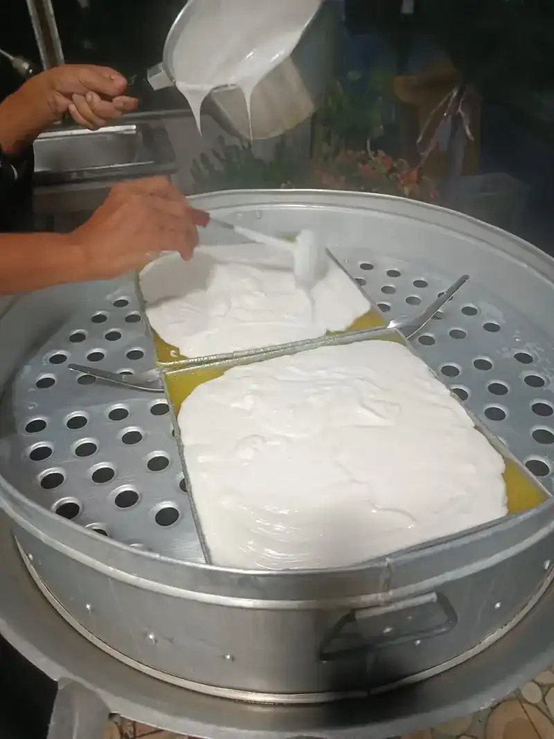 Hands pouring a white top layer over trays in a large steamer