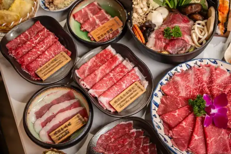 Plates of marbled beef cuts and a hotpot of vegetables and mushrooms, seen from above