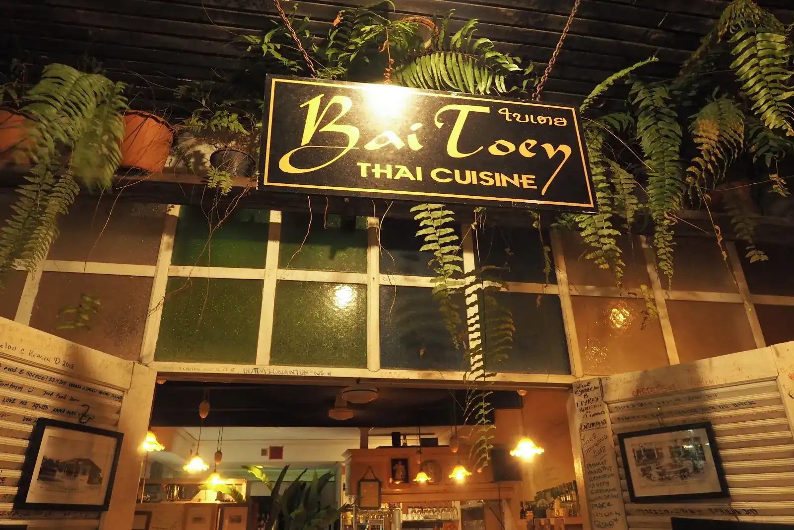 The Bai Toey sign, gold letters on black, under hanging ferns at the front of the restaurant at night