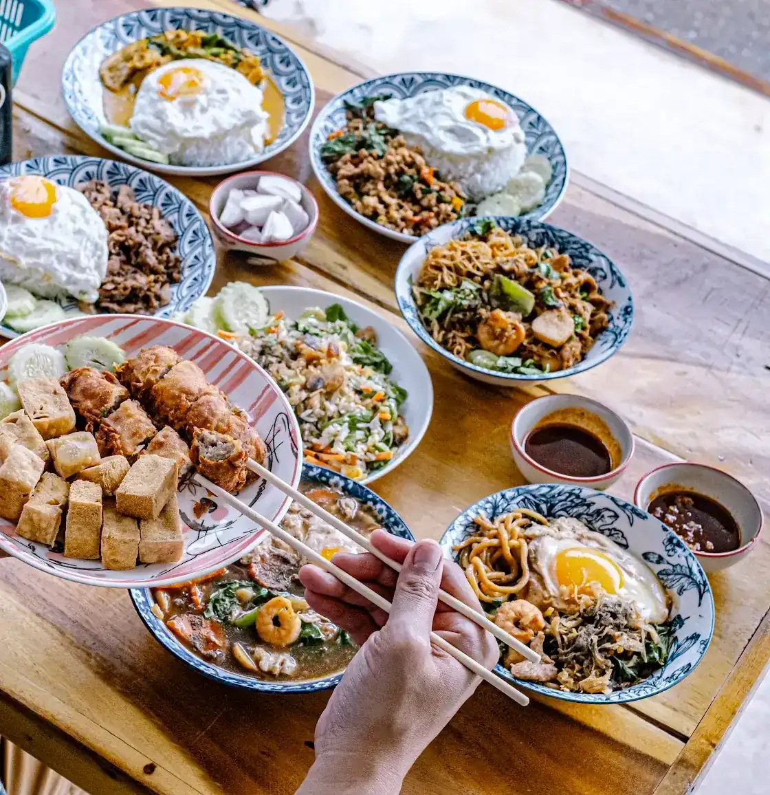A table of dishes: fried tofu and sliced fried rolls held up on a plate, rice with fried eggs, noodle bowls and dipping sauces