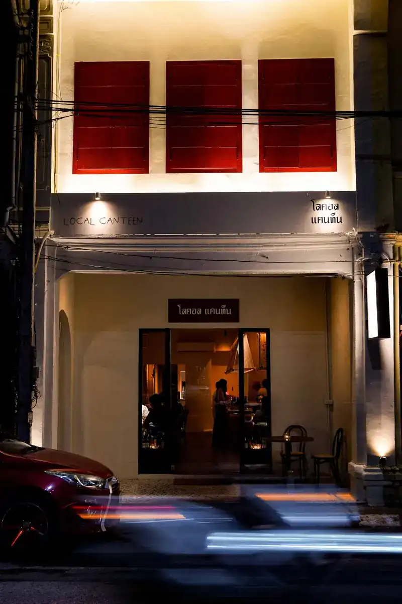 The shophouse front at night, red shutters above the lit doorway