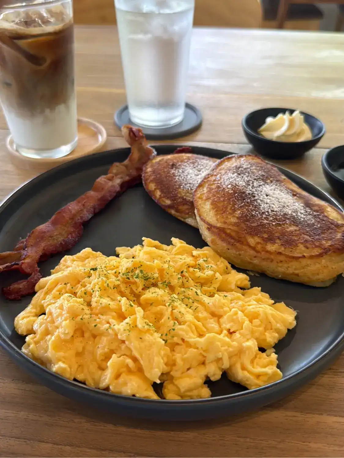 Scrambled eggs, two pancakes and bacon on a dark plate, with an iced coffee and a glass of water