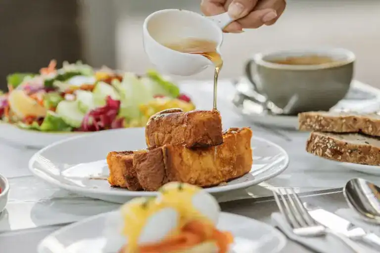 Syrup being poured over French toast on a breakfast table with salad, coffee and seeded bread