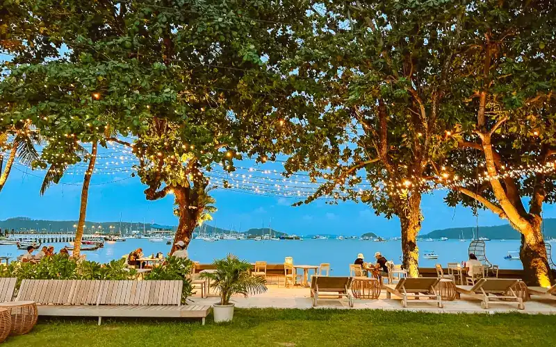 The garden at dusk: strings of lights in the trees, sun loungers on the lawn and boats on Chalong Bay