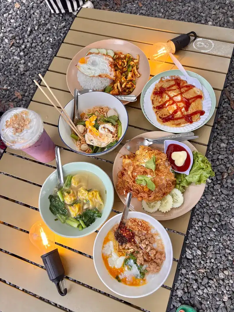 From above: rice with a fried egg, a crispy omelette, wonton soup, rice porridge, noodles and a pink iced drink on a slatted camp table lit by two lanterns