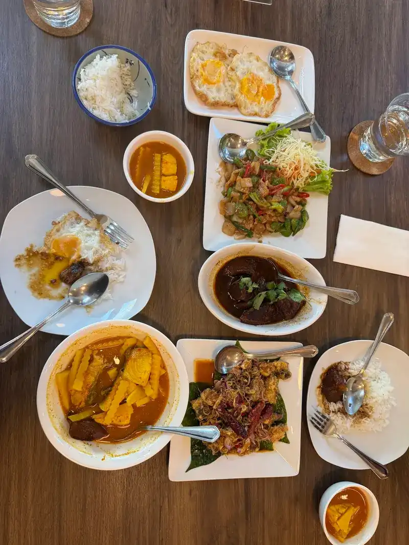 Shared dishes on a wooden table, seen from above: a yellow curry, moo hong, stir-fries, fried eggs and rice