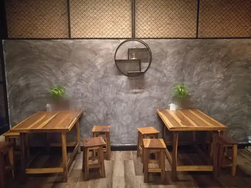 Two wooden tables with stools in front of a polished concrete wall, chain-link panels above and a round iron shelf