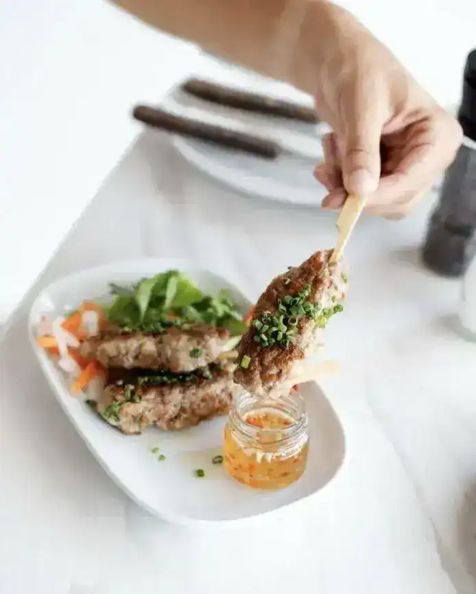A skewer lifted from a plate of pickled vegetables and herbs, with dipping sauce