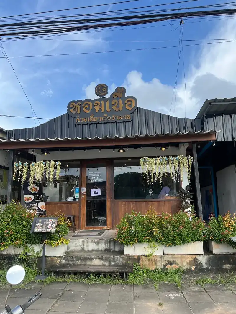 The shop front: a dark roof with the gold หอมเนื้อ sign, wooden doors and windows, and plants along the front
