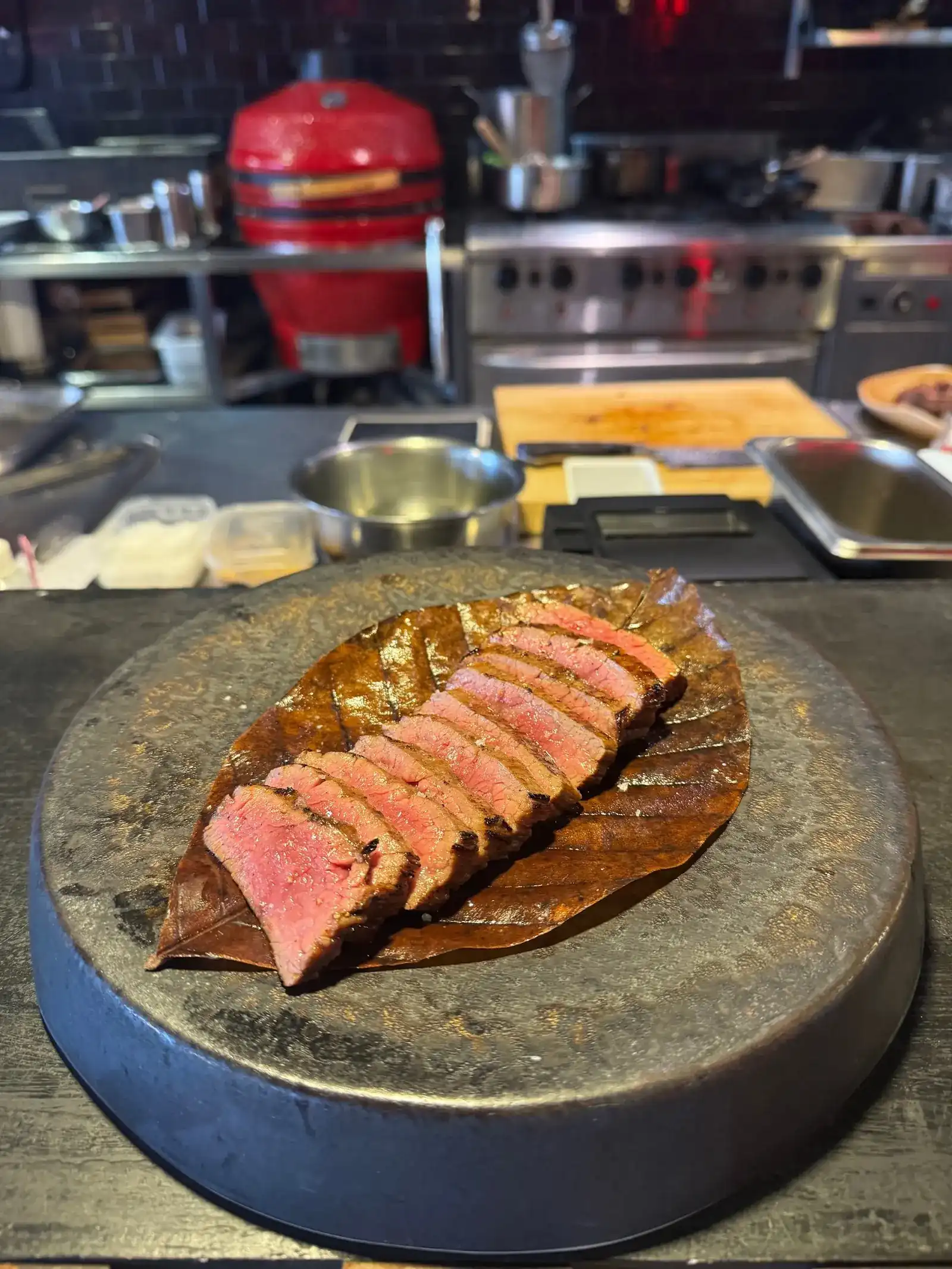 Sliced grilled beef on a leaf, with the red charcoal grill of the open kitchen behind / เนื้อย่างหั่นชิ้นวางบนใบไม้ ด้านหลังเป็นเตาถ่านสีแดงในครัวเปิด