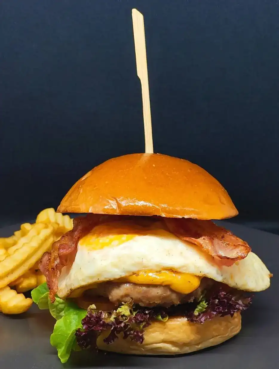 Burger with a fried egg, bacon, cheese and lettuce in a glossy bun, crinkle-cut fries beside it