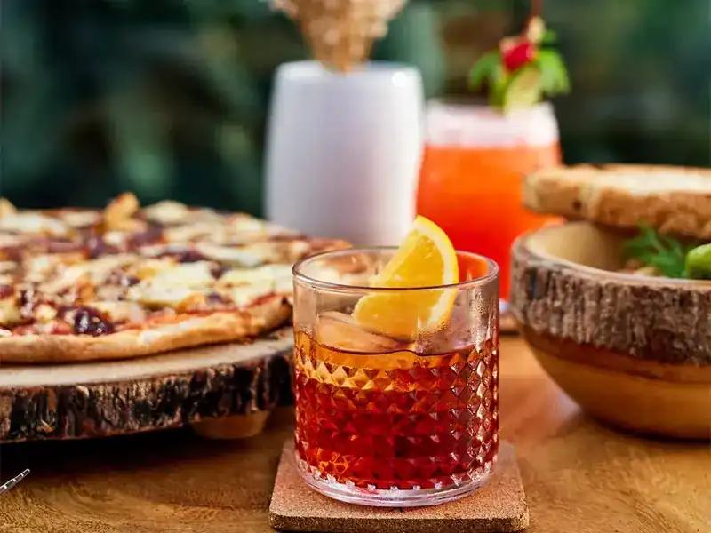Pizza on a slice of tree trunk beside two mixed drinks