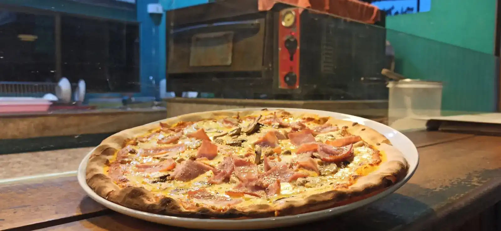 Ham and mushroom pizza on the counter, the pizza oven behind it