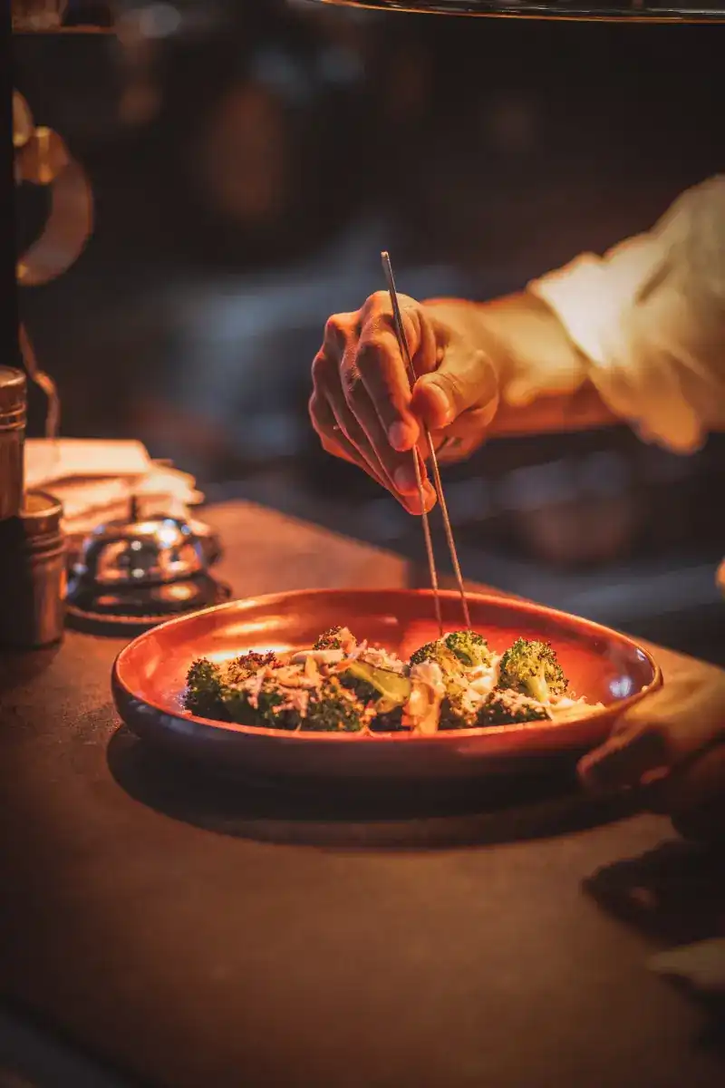A chef's hand finishes a plate with long tweezers at the pass