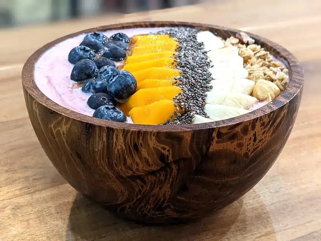 Smoothie bowl in a wooden bowl, topped with blueberries, mango, chia seeds, banana and granola