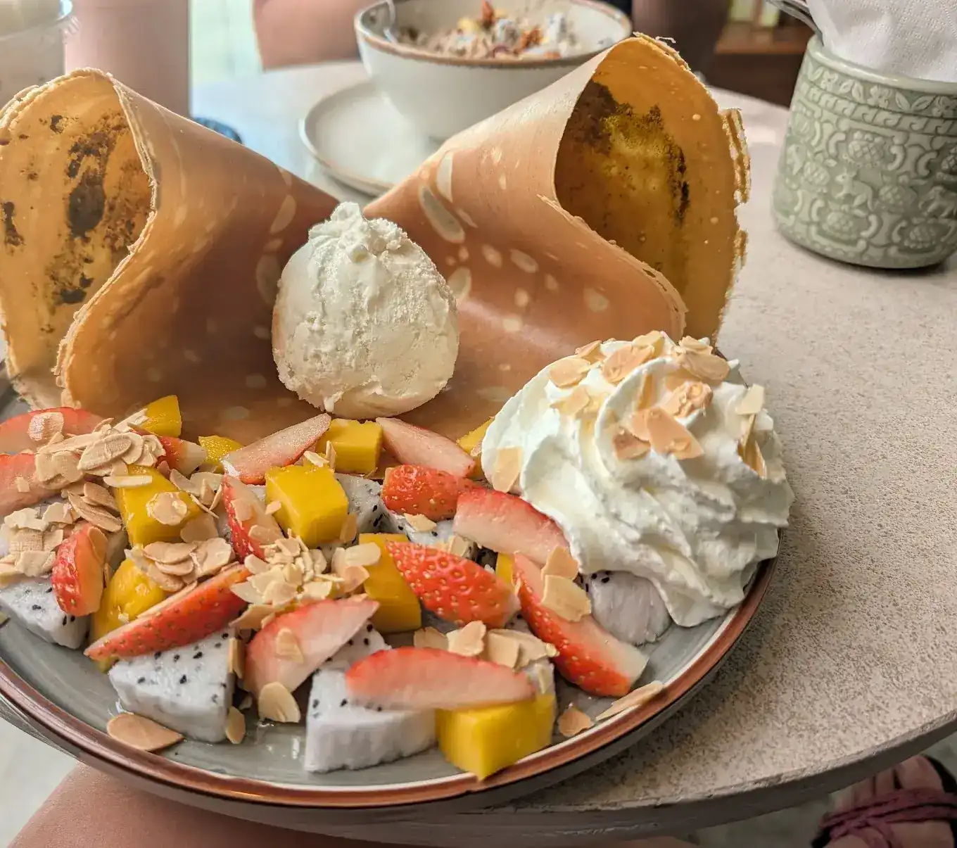 A sweet crêpe folded upright around a scoop of ice cream, with strawberries, mango, dragon fruit, whipped cream and toasted almonds