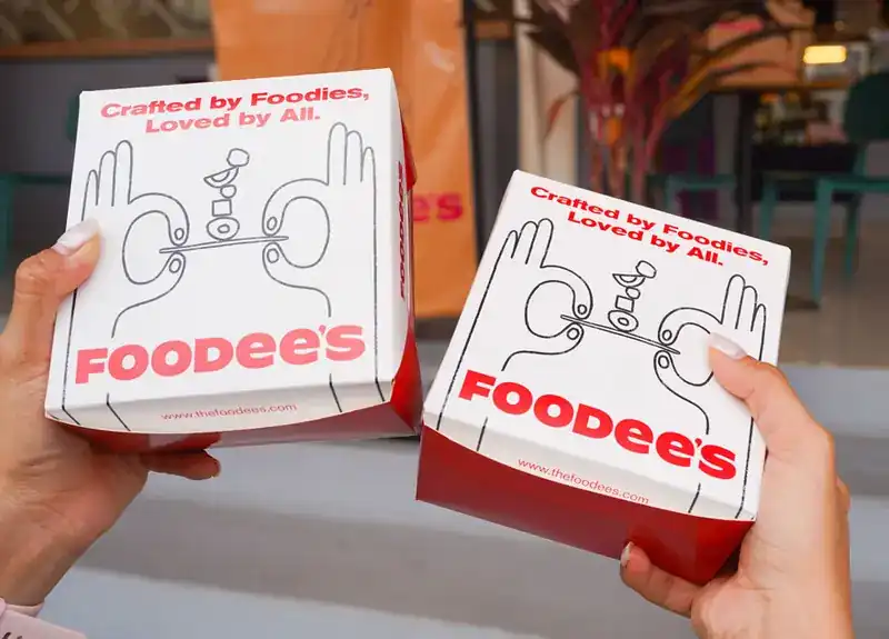 Two hands holding red and white FOODEE’S takeaway boxes