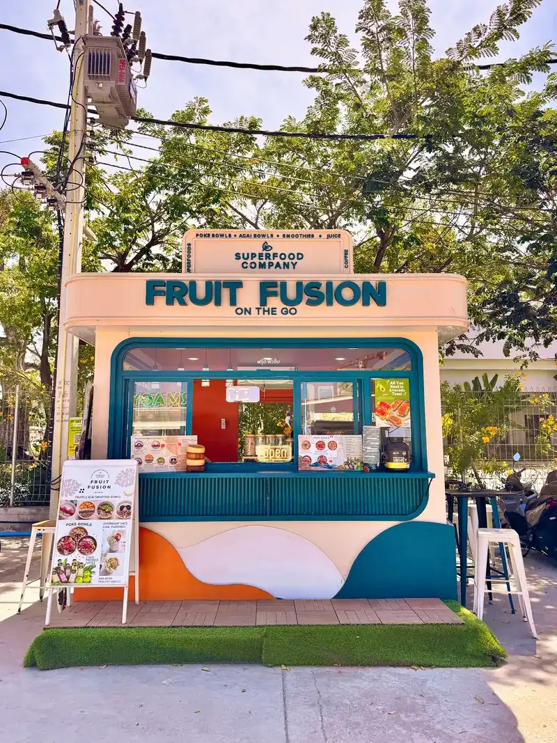 The Fruit Fusion On the Go kiosk: a cream booth with a teal window, an orange-and-white wave painted on its base and white stools on artificial grass