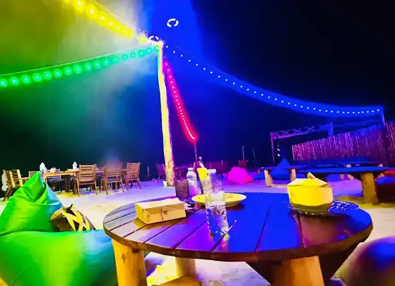 Coloured light strings over tables and beanbags on the sand at night