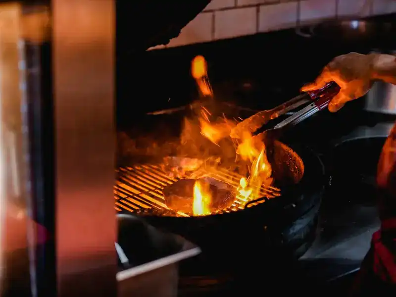 Flames rising through the grill as a cook turns the meat with tongs