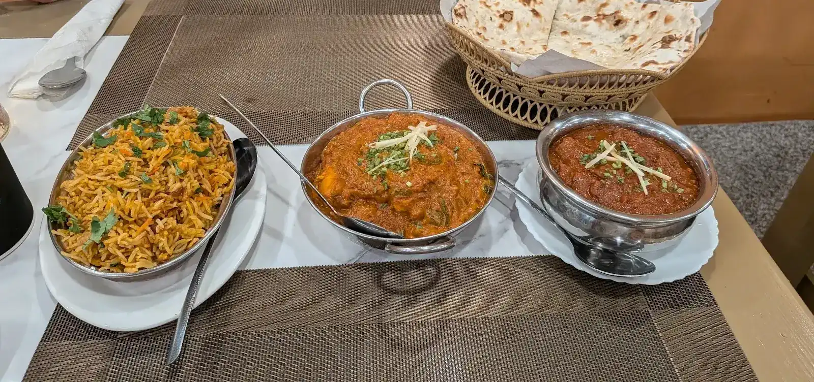 Spiced rice, two curries in steel bowls and a basket of naan on a table