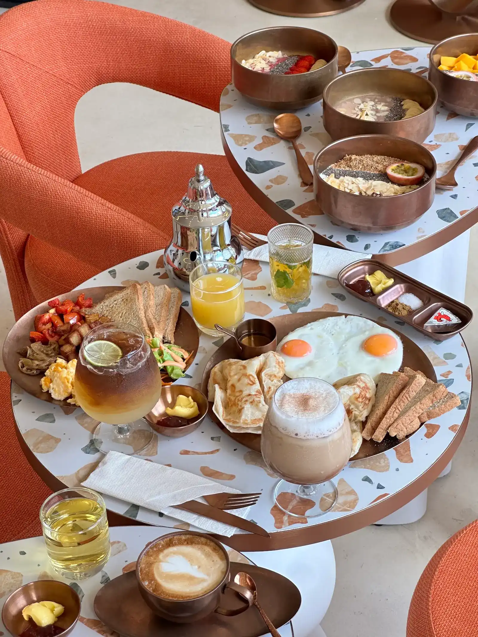 Breakfast for a table: fried eggs, flatbreads, toast, juice and mint tea on a terrazzo table, with smoothie bowls behind