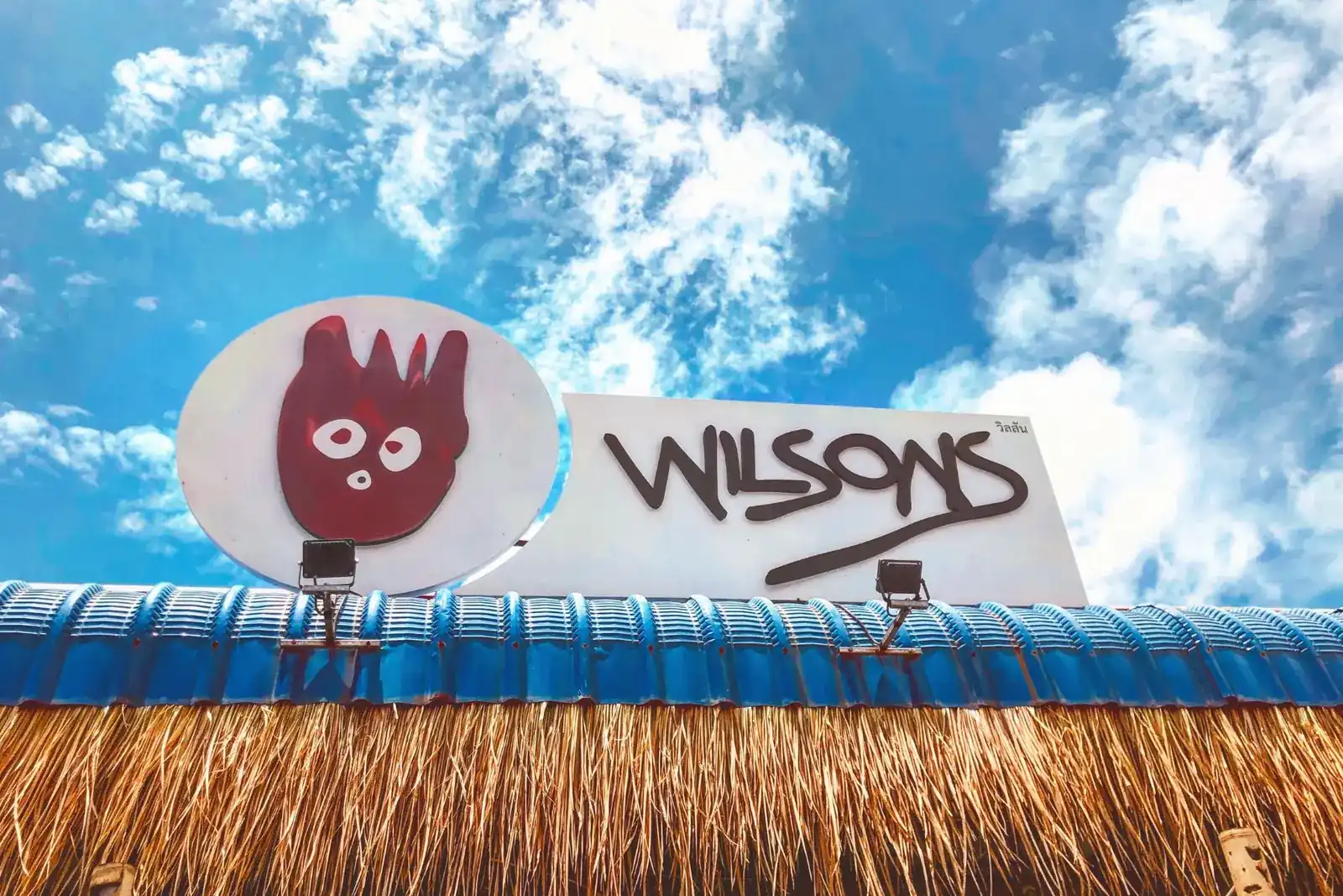 The Wilsons sign with its red handprint face on the thatched roof, against a blue Rawai sky