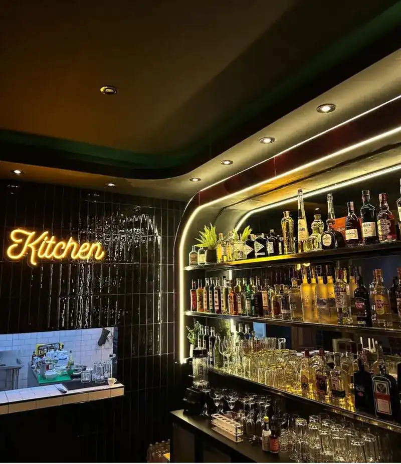 Yellow neon sign reading Kitchen on black tiles, above the kitchen pass