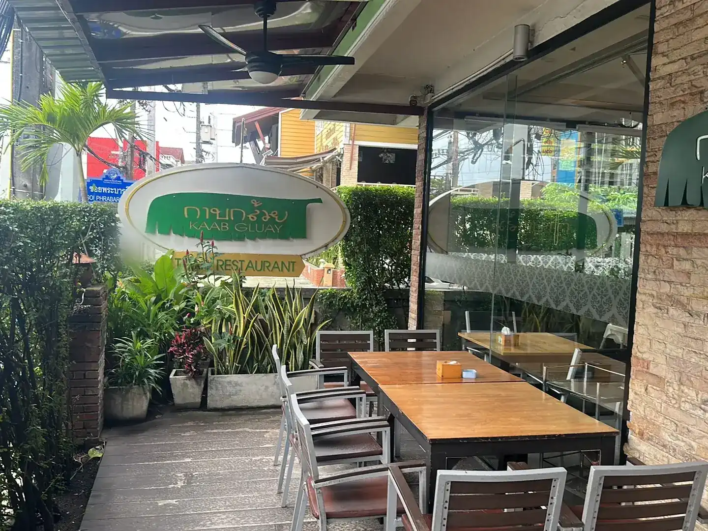 The planted front terrace of Kaab Gluay on Phrabaramee Road, wooden tables under the green and white sign