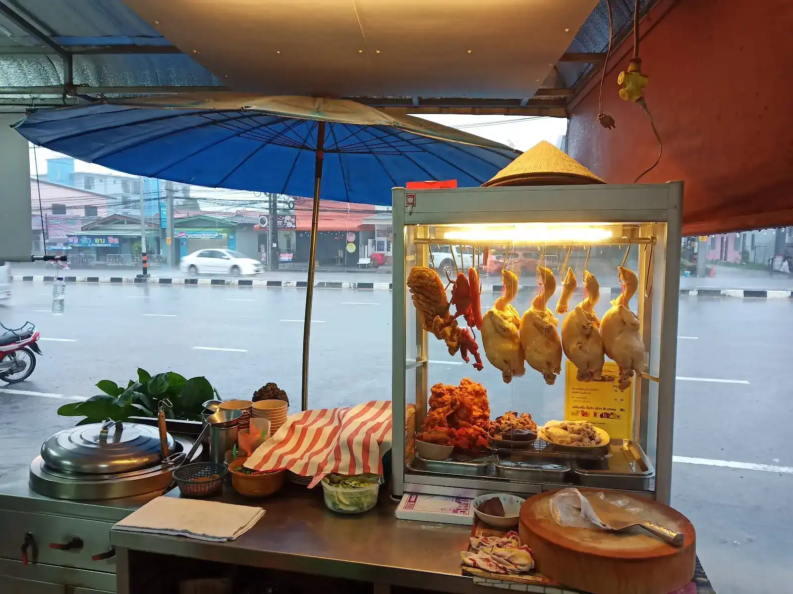 The lit glass case of whole chickens under a blue umbrella, by the road on a rainy day
