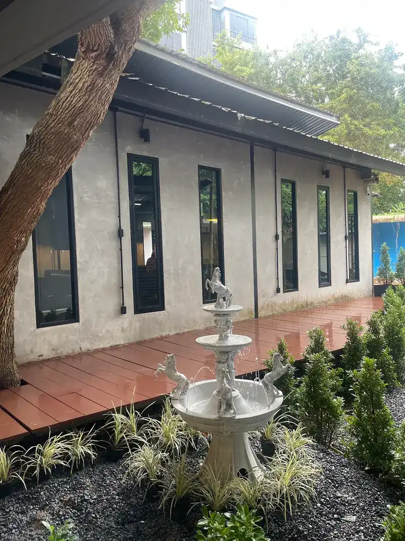 A wooden deck along the building's tall windows, with a white tiered fountain in a garden bed and a tree trunk beside it