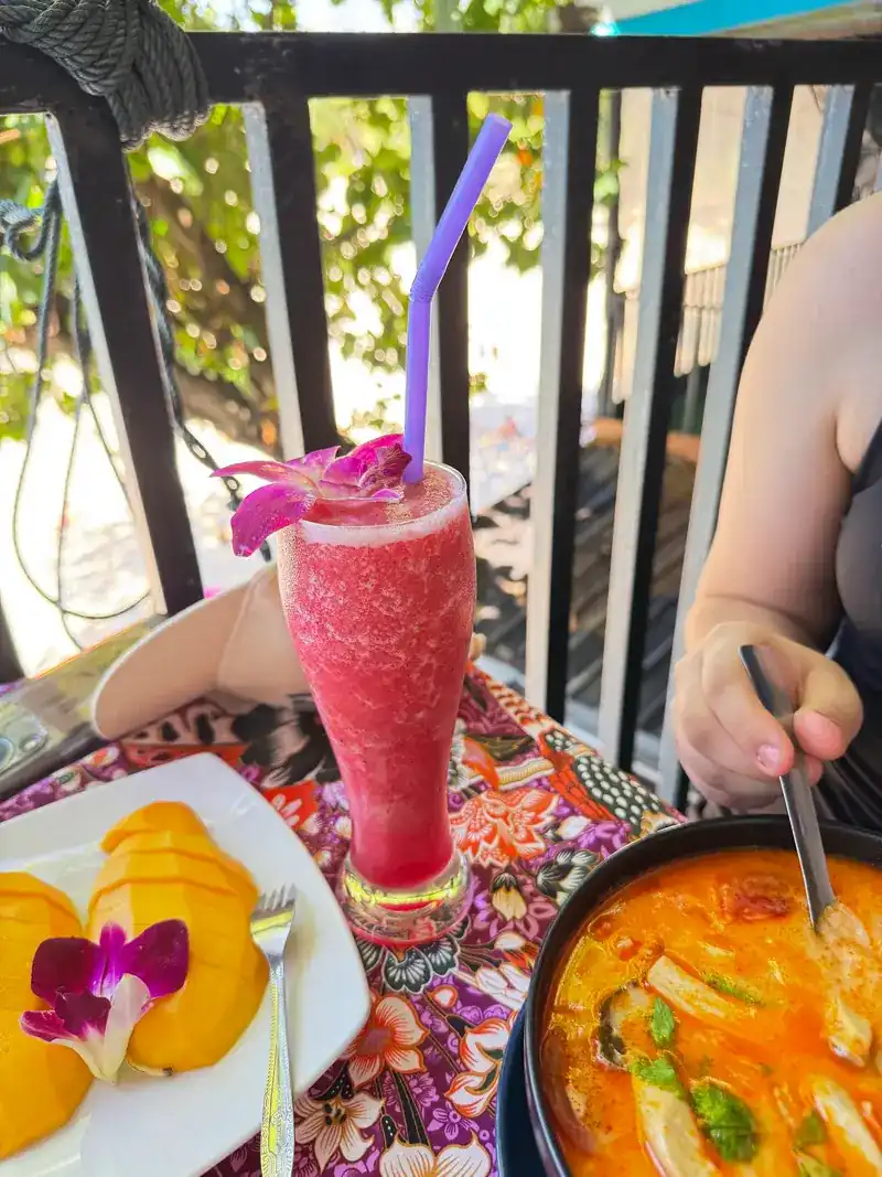 A bowl of orange soup, sliced mango with an orchid and a red fruit shake by the railing.
