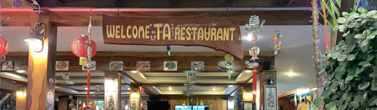 The carved wooden sign Welcome..Ta Restaurant over the dining room, with red lanterns / ป้ายไม้แกะสลัก Welcome..Ta Restaurant เหนือห้องอาหาร ประดับโคมแดง