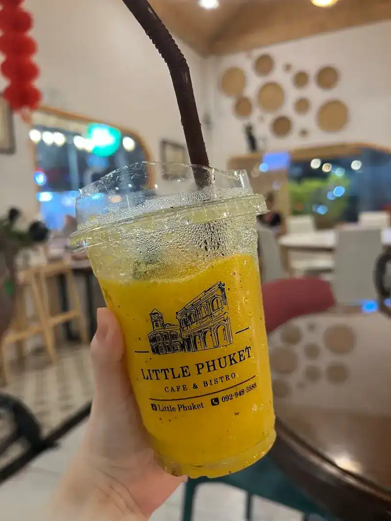 A yellow fruit smoothie in the printed Little Phuket cup, held up inside the restaurant