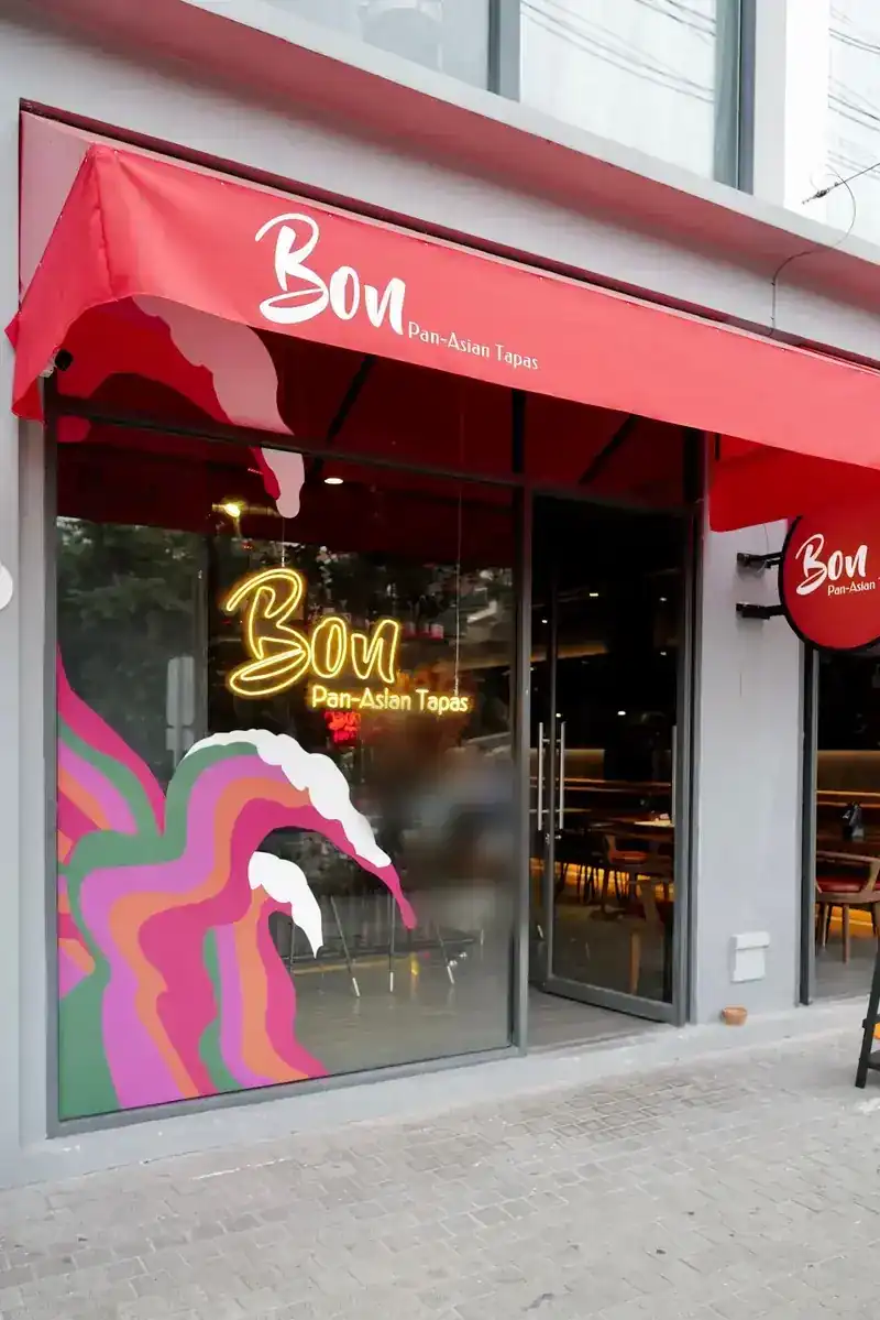 The restaurant front: a red awning, a yellow neon Bon sign and a colourful wave painted on the window / หน้าร้าน กันสาดสีแดง ป้ายไฟนีออน Bon สีเหลือง และภาพคลื่นหลากสีบนกระจก