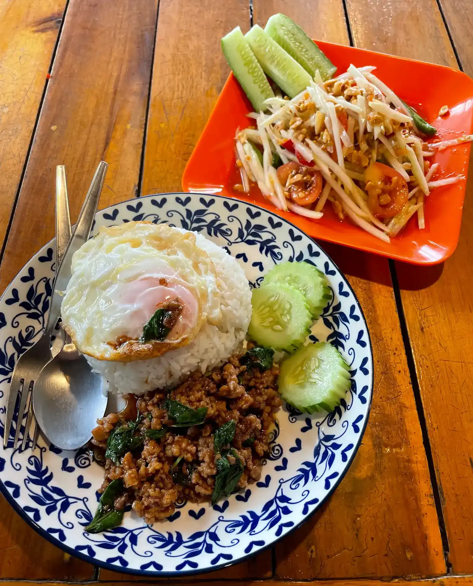 Stir-fried basil pork with a fried egg on rice, and a papaya salad on an orange plate / ผัดกะเพราหมูไข่ดาวราดข้าว กับส้มตำในจานสีส้ม