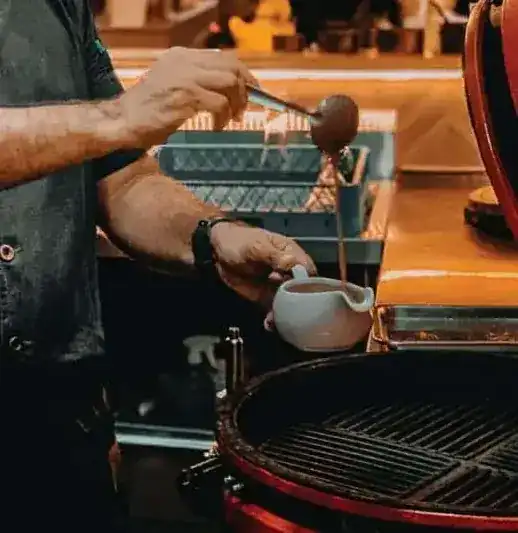 A cook's hands ladle sauce into a small white jug beside a charcoal grill