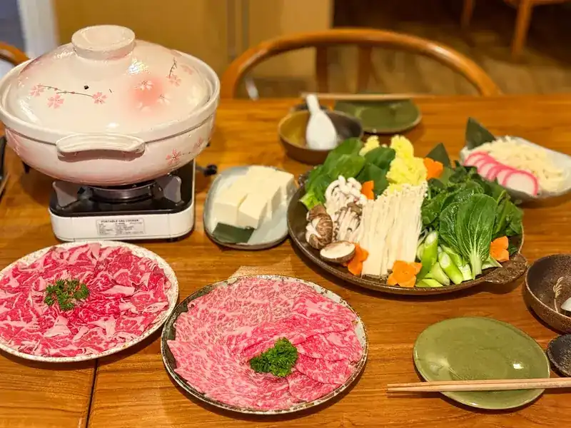 A clay hot pot on a burner with plates of sliced marbled beef, vegetables, mushrooms and tofu