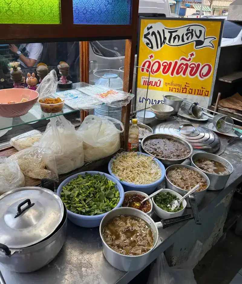 The cart: noodles in bags, morning glory, bean sprouts, chilli, chopped herbs and pots of broth and braised beef, with the yellow shop sign behind / รถเข็นหน้าร้าน มีเส้น ผักบุ้ง ถั่วงอก พริก ผักชีต้นหอม และหม้อน้ำซุปกับเนื้อตุ๋น ป้ายร้านสีเหลืองด้านหลัง
