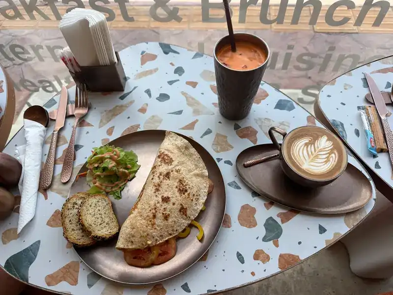 A folded flatbread with salad and bread, a latte and a cold drink by the front window
