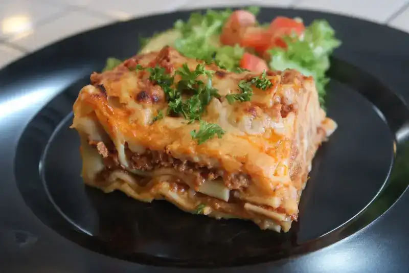 A slice of baked lasagne with salad on a black plate