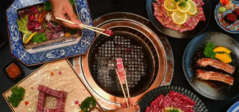 Seen from above: chopsticks lay strips of marbled beef on a copper-rimmed charcoal grill, with plates of raw cuts around it