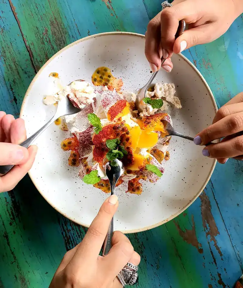 Four hands with spoons sharing a pavlova with mango and passion fruit