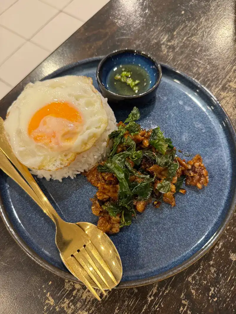 Holy basil stir-fry on rice with a fried egg and a small bowl of chilli fish sauce, on a blue plate