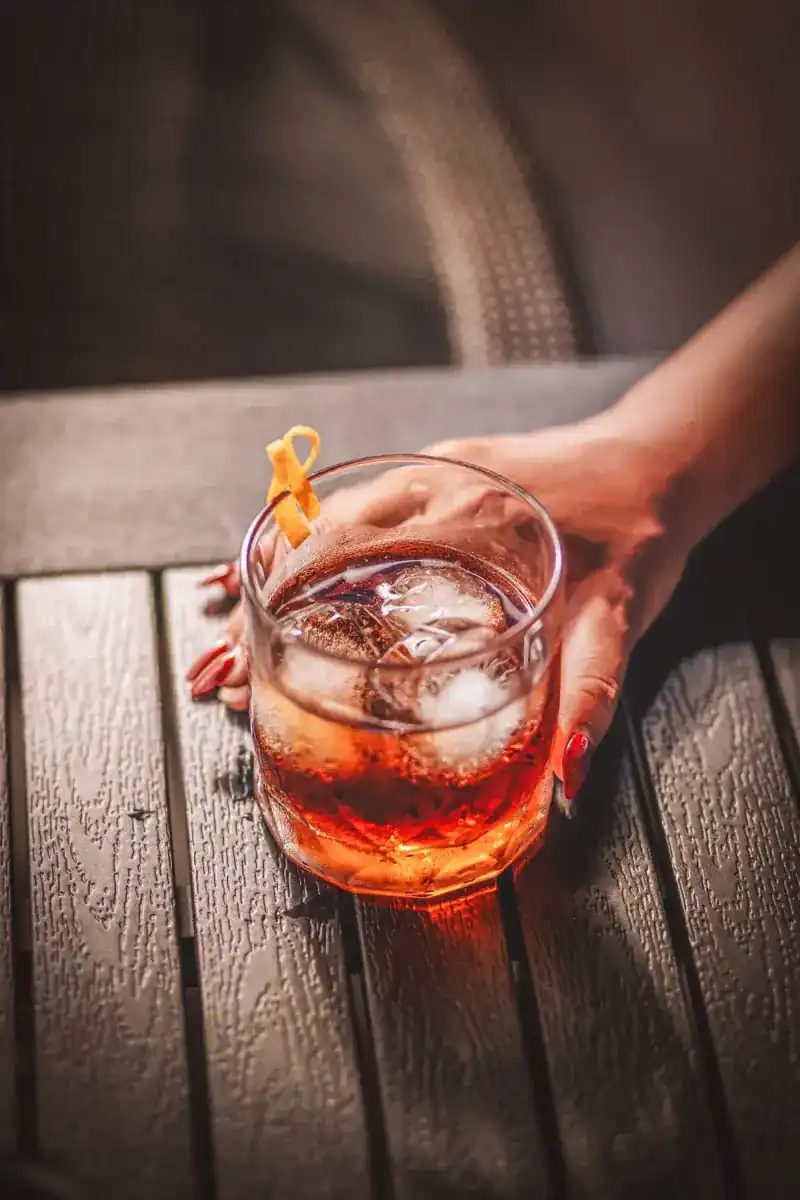 A hand rests on a short drink over ice with a twist of orange peel, on a slatted table