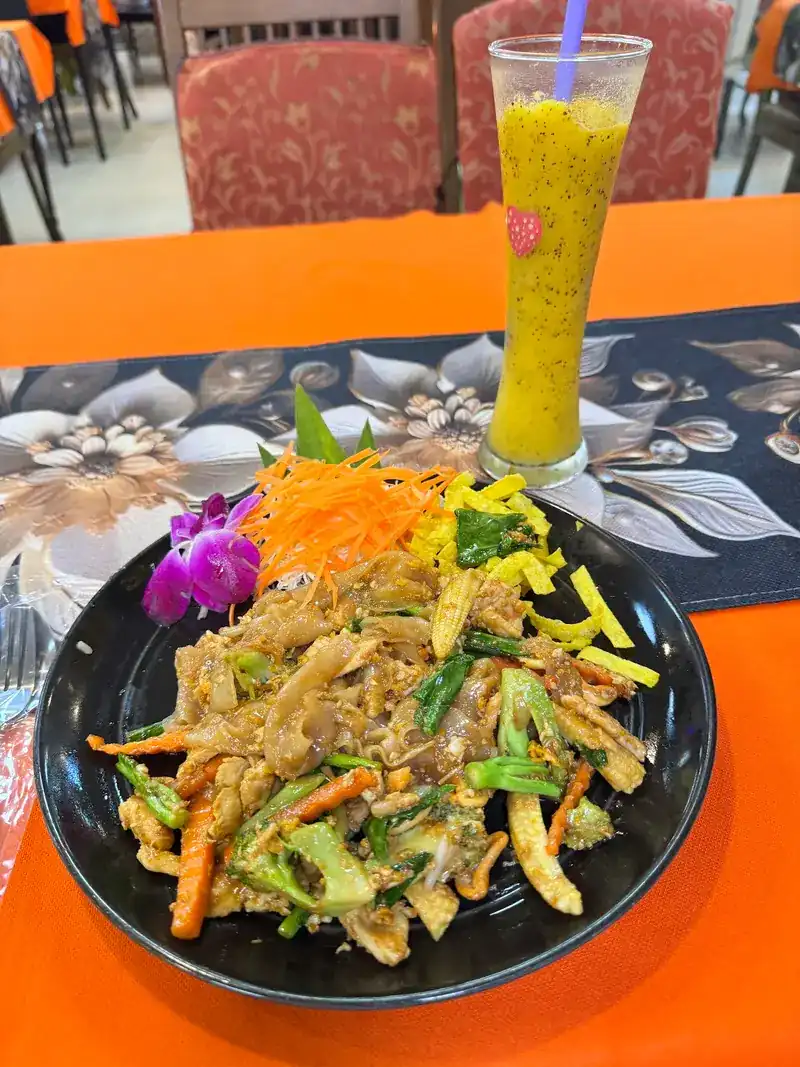 Stir-fried rice noodles with vegetables and an orchid, and a yellow fruit shake, on an orange tablecloth