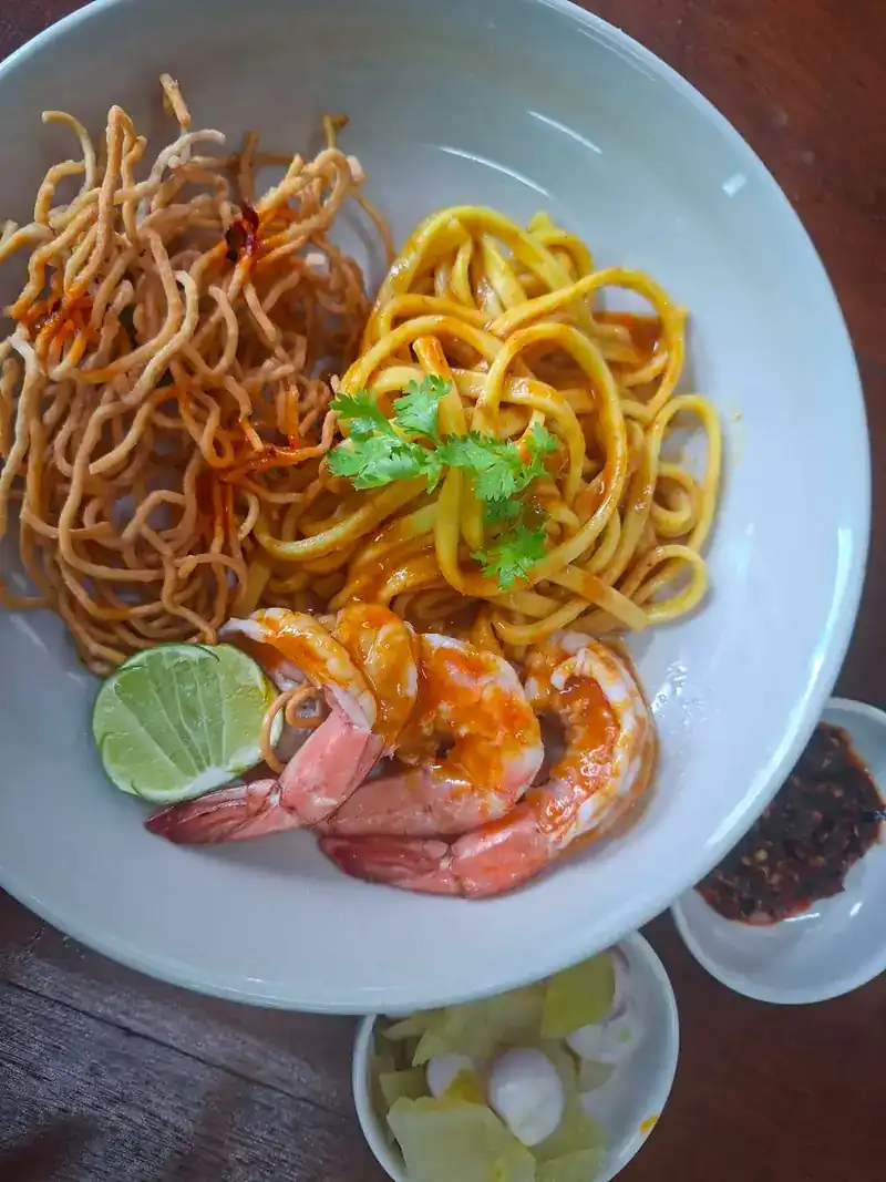 Khao soi seen from above: yellow egg noodles, crisp fried noodles, three prawns and a slice of lime, with chilli paste, shallots and pickled greens in small bowls